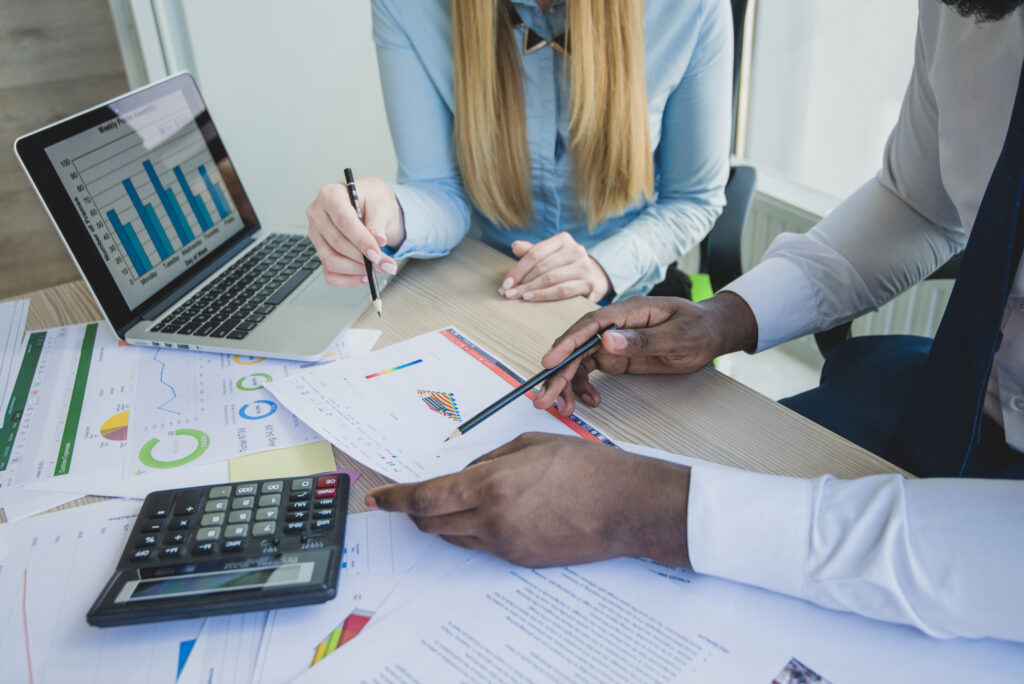 Duas pessoas analisando documentos financeiros em uma mesa. Há gráficos coloridos em papéis e na tela de um notebook, uma calculadora, e ambos apontam com canetas para os relatórios. A cena representa um ambiente de trabalho corporativo focado em análise de dados ou contabilidade.