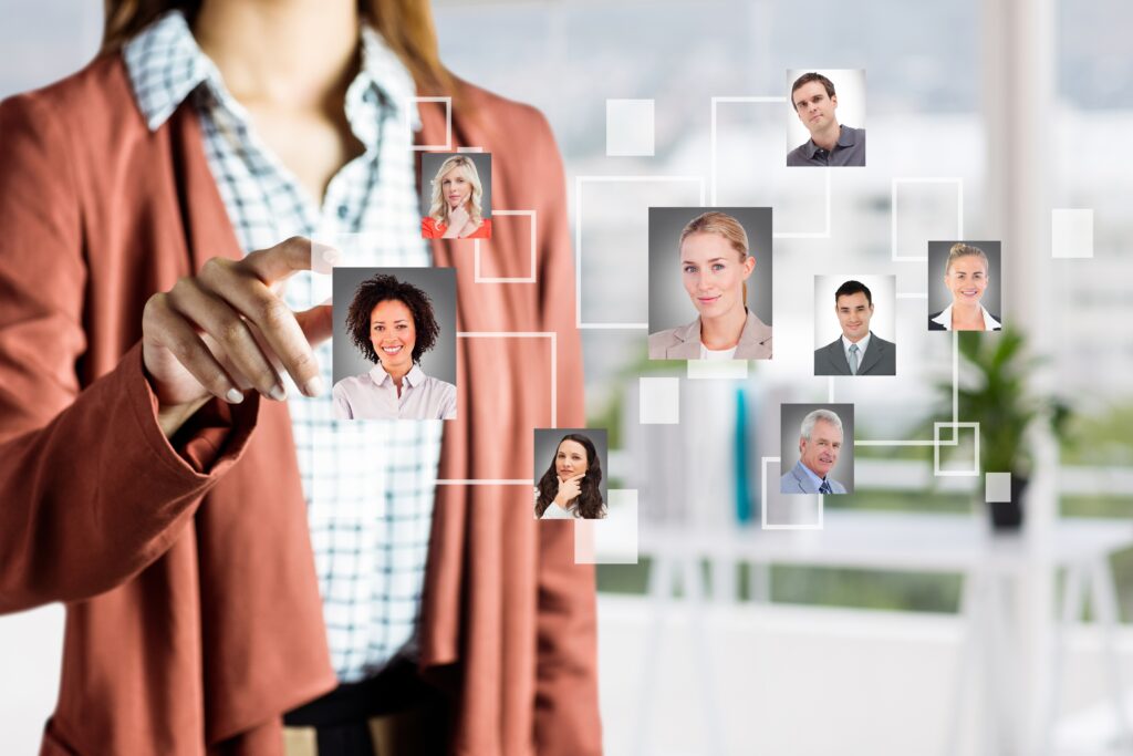 Mulher interagindo com uma interface digital que mostra uma rede de perfis profissionais, simbolizando conexão e gestão de equipe. Ela toca a imagem de uma mulher sorridente, enquanto outros rostos aparecem conectados por linhas e quadrados em um fundo de escritório moderno.