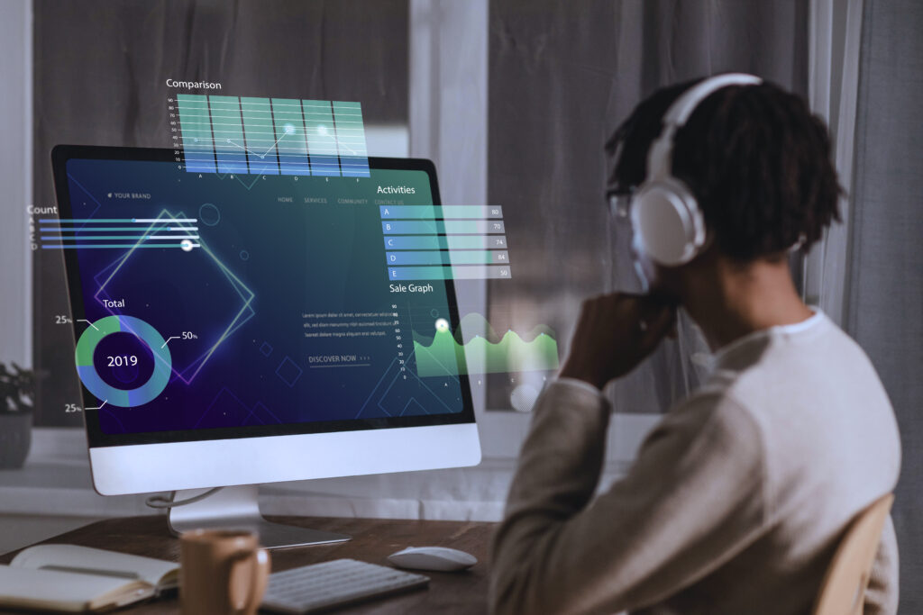 Pessoa com fones de ouvido observando um monitor de computador com gráficos analíticos flutuantes na tela, incluindo gráficos de comparação, atividades, vendas e um gráfico de rosca com o ano de 2019 destacado. A cena sugere análise de dados ou trabalho com visualização de informações em ambiente moderno e tecnológico.